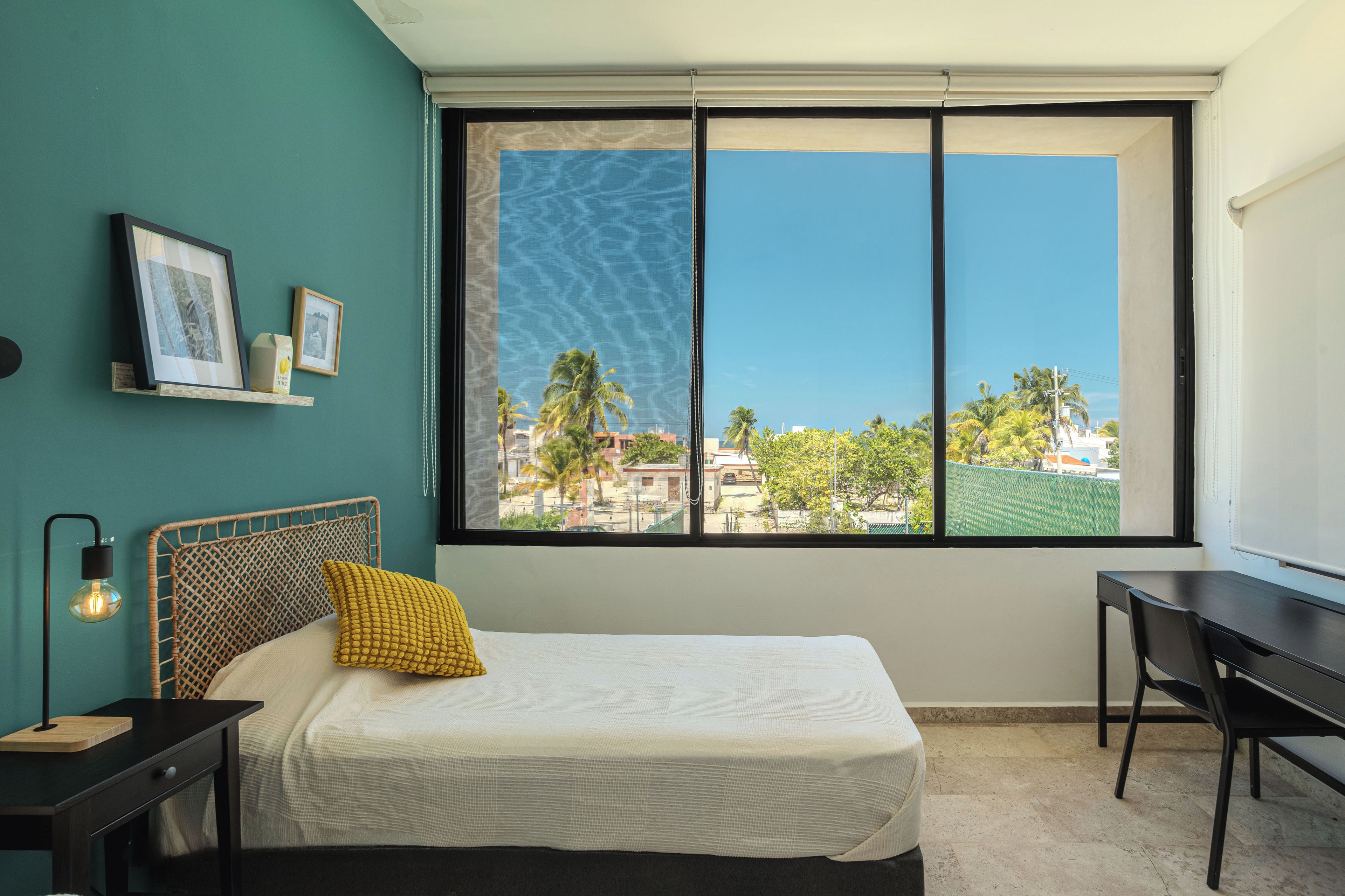 A bedroom with a bed, a desk, and a window with a view of palm trees and buildings.