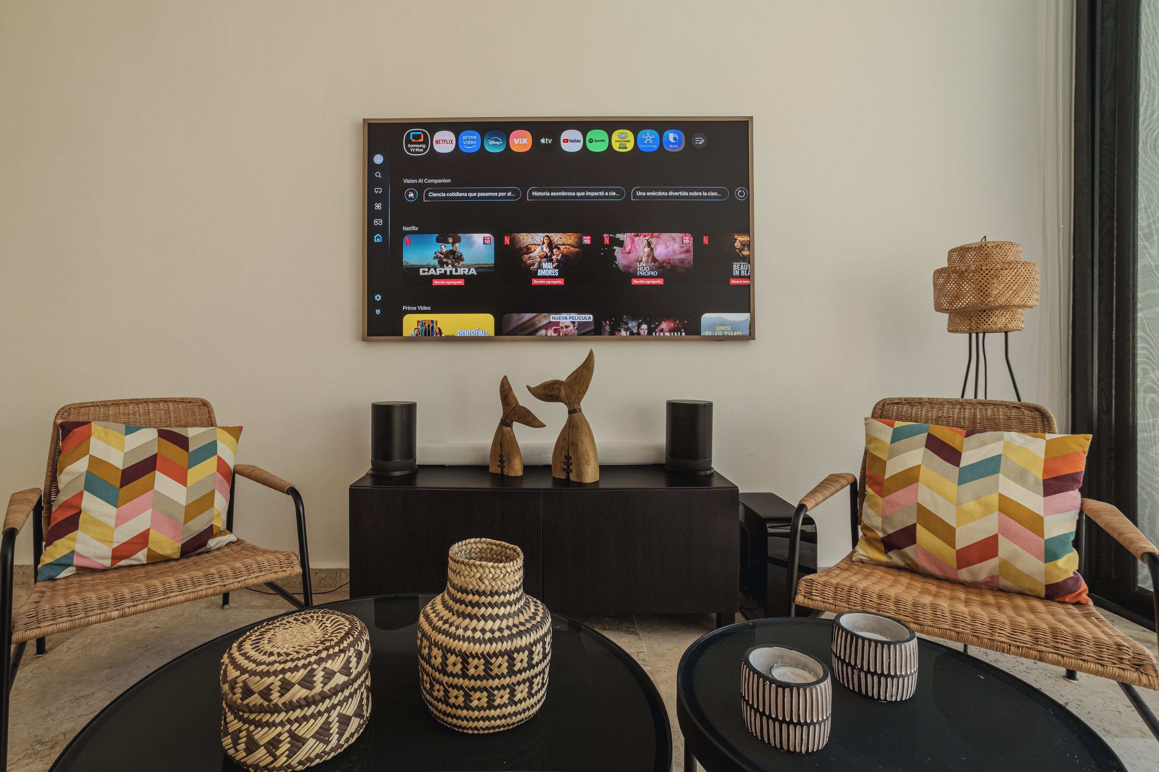 A living room with a large flat-screen TV mounted on the wall displaying various streaming services. There are two wicker chairs with colorful cushions, a black coffee table with woven baskets, and a small side table with a lamp.