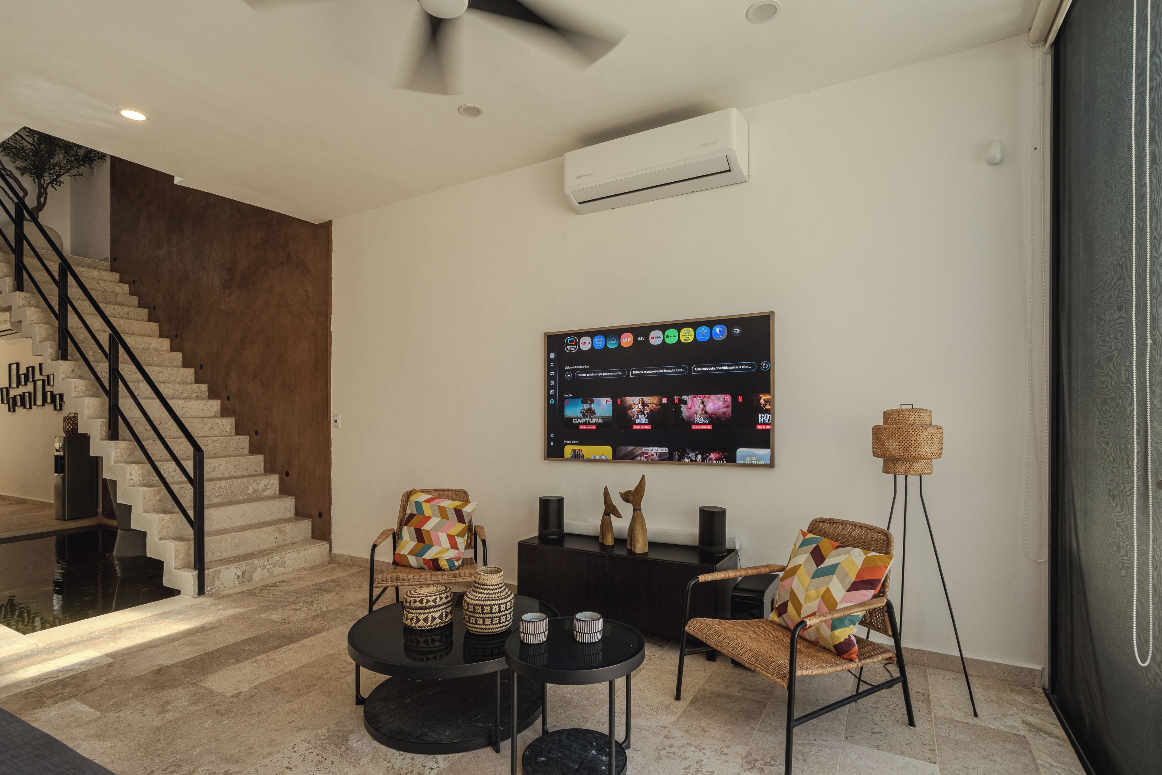 A modern living room with a staircase, a flat-screen TV mounted on the wall, and a coffee table with decorative items.