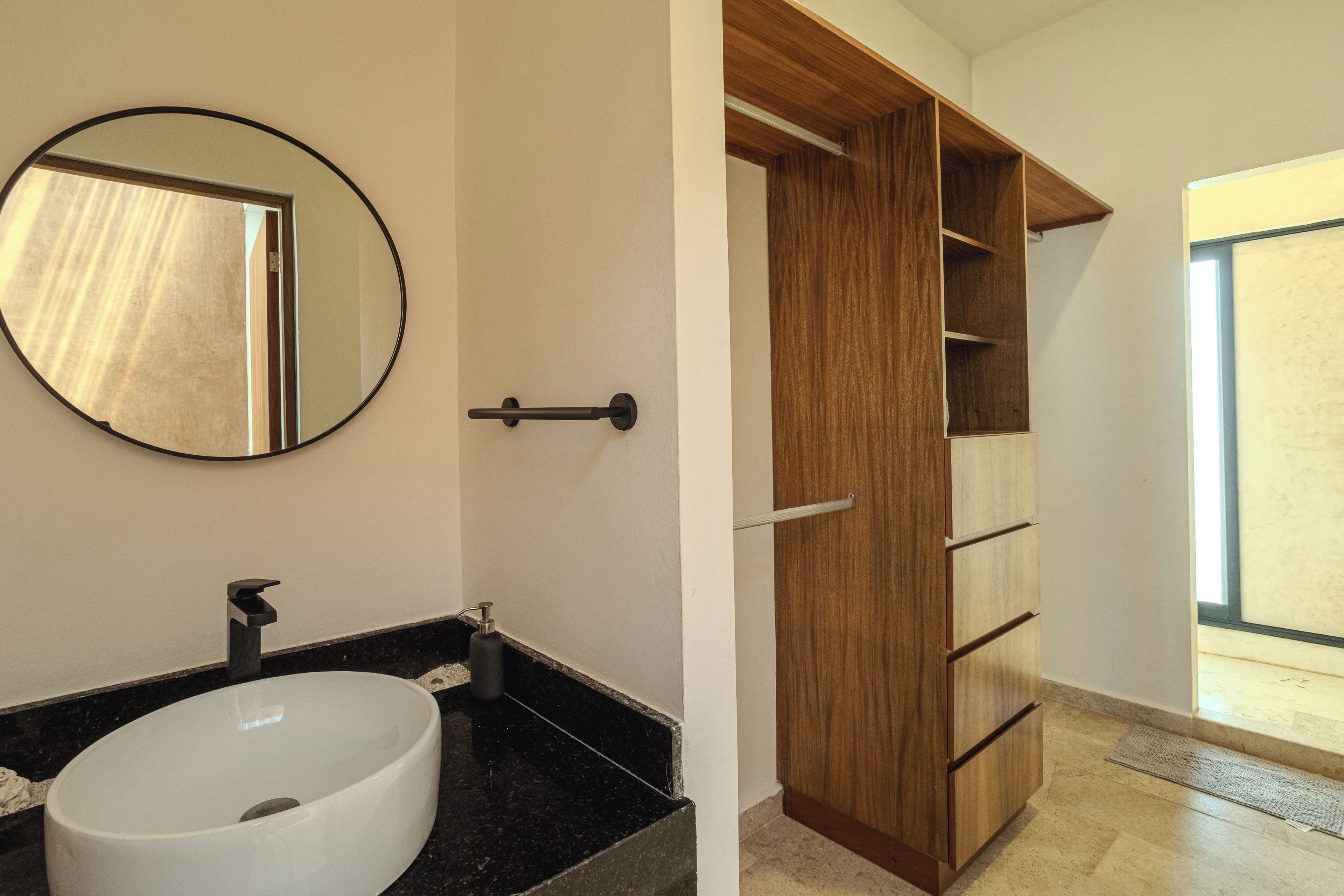 A bathroom with a round mirror, a white sink, and a wooden wardrobe with multiple shelves.