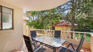 A balcony with a glass table and chairs, surrounded by a railing, overlooking a garden with trees and a building with a red roof. - Lovely Mermaid Beach Apartment (Gold Coast)