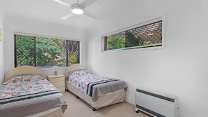 A bedroom with two single beds, a bedside table, a window with a view of greenery, and a ceiling fan. - Lovely Mermaid Beach Apartment (Gold Coast)