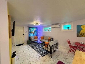 A living room with a sofa, a coffee table, and a TV. There is a door leading to another room and a window with a view of trees.