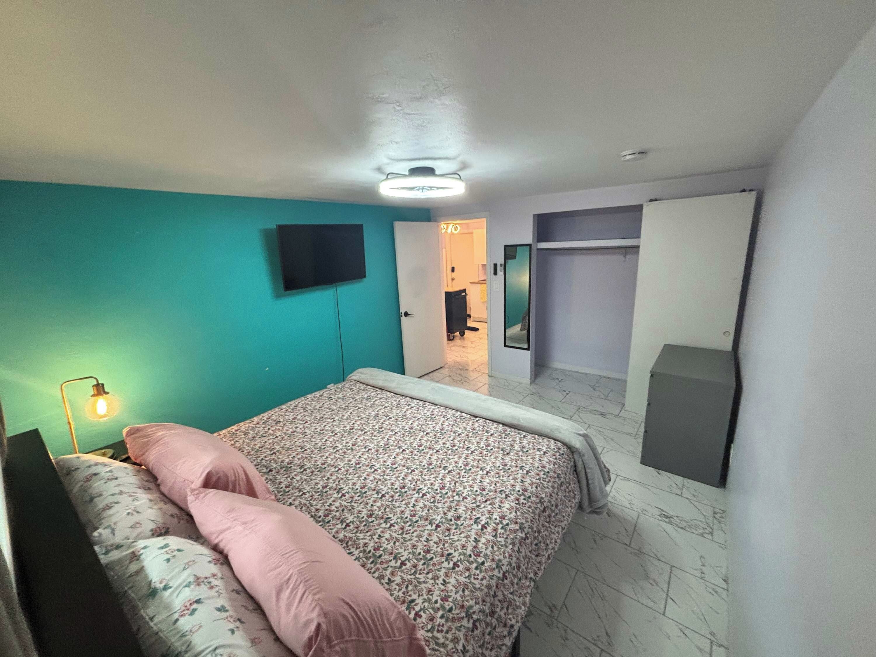Une chambre à coucher avec un lit, un téléviseur fixé au mur, une armoire et une porte donnant sur une autre pièce.