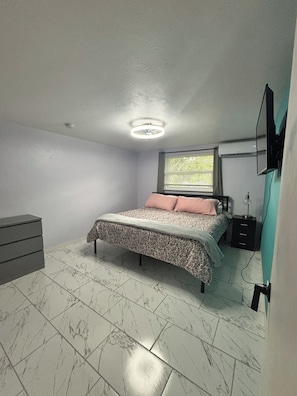 A bedroom with a bed, a dresser, a TV mounted on the wall, and a window with blinds.