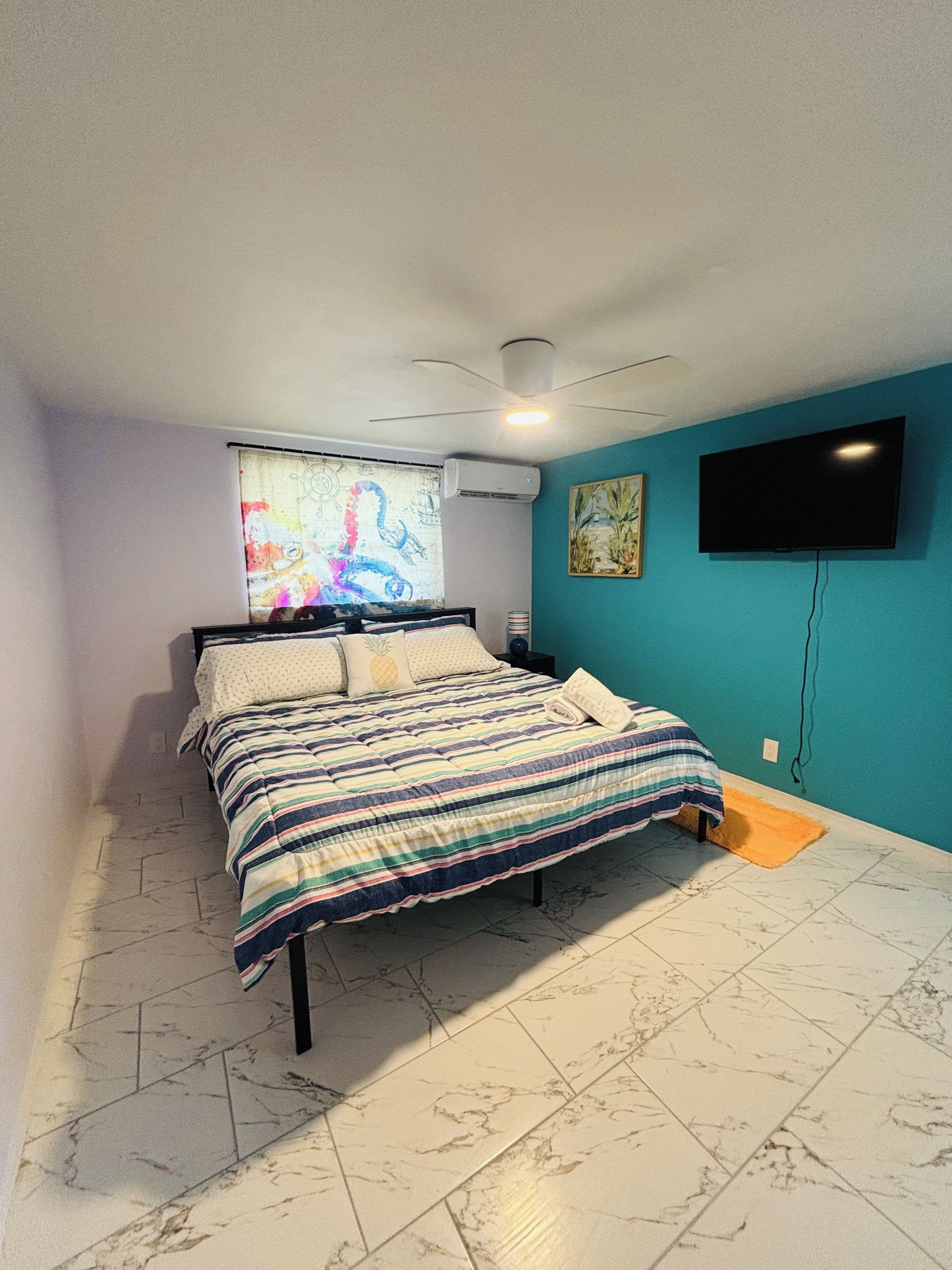 A bedroom with a bed, a ceiling fan, an air conditioning unit, a television on the wall, and a window with a colorful curtain.