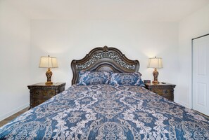 A bed with a patterned bedspread, flanked by two nightstands with lamps, in a room with plain walls and a door. - 100" TV - Pool, Spa & Epic Game Room Villa for 14! (Kissimmee)