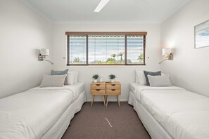 Ein Schlafzimmer mit zwei Einzelbetten, einem kleinen Holfnachtkästchen mit zwei Blumentöpfen und einem Fenster mit Jalousien.