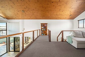 Ein geräumiger Raum mit Holzbalkendecke, einer eingebauten Bücherwand, einem Sofa und Blick durch eine Glastür auf einen Außenbereich.