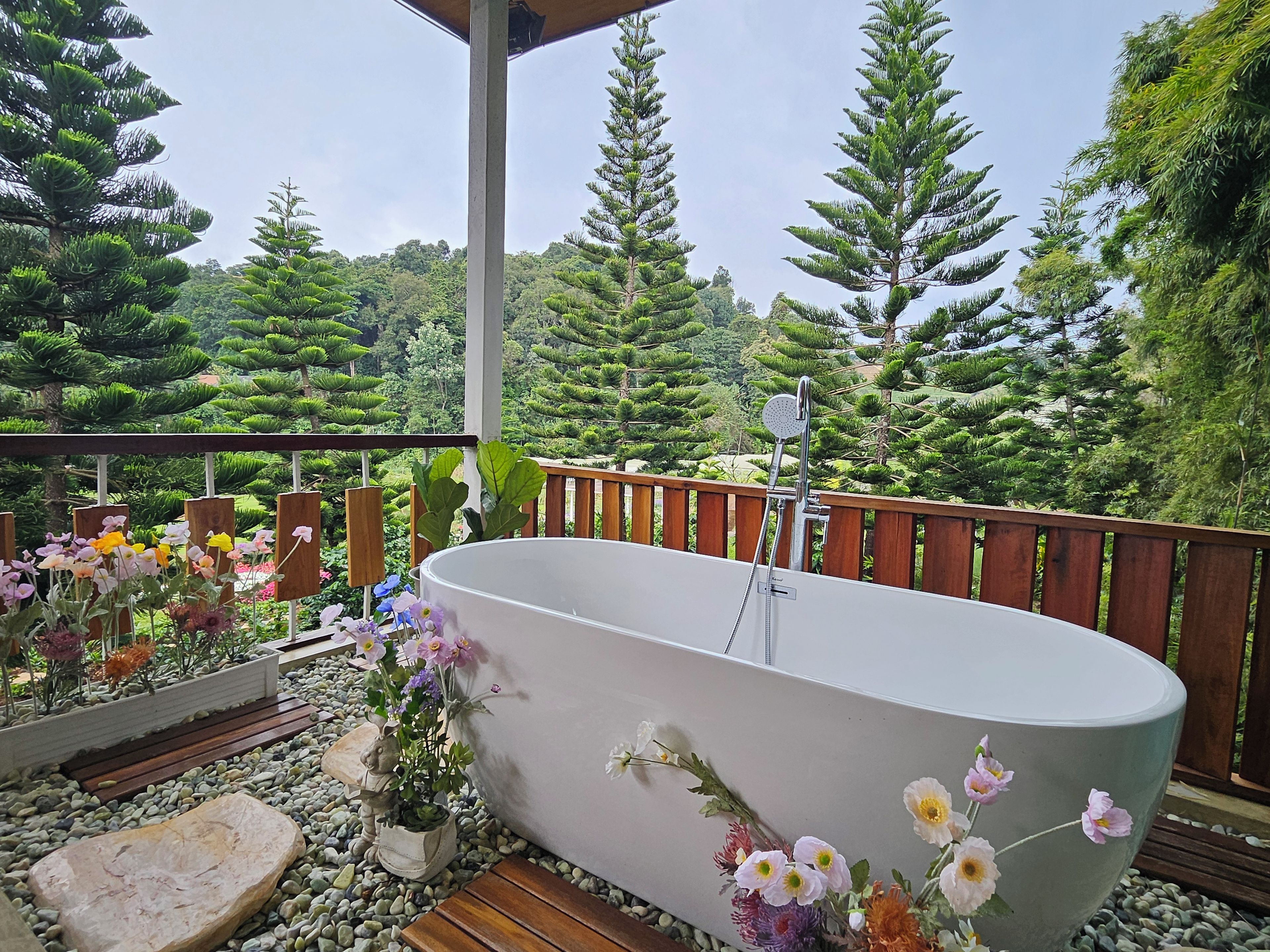 A freestanding bathtub with a showerhead, surrounded by a wooden deck, with a view of a forested area.