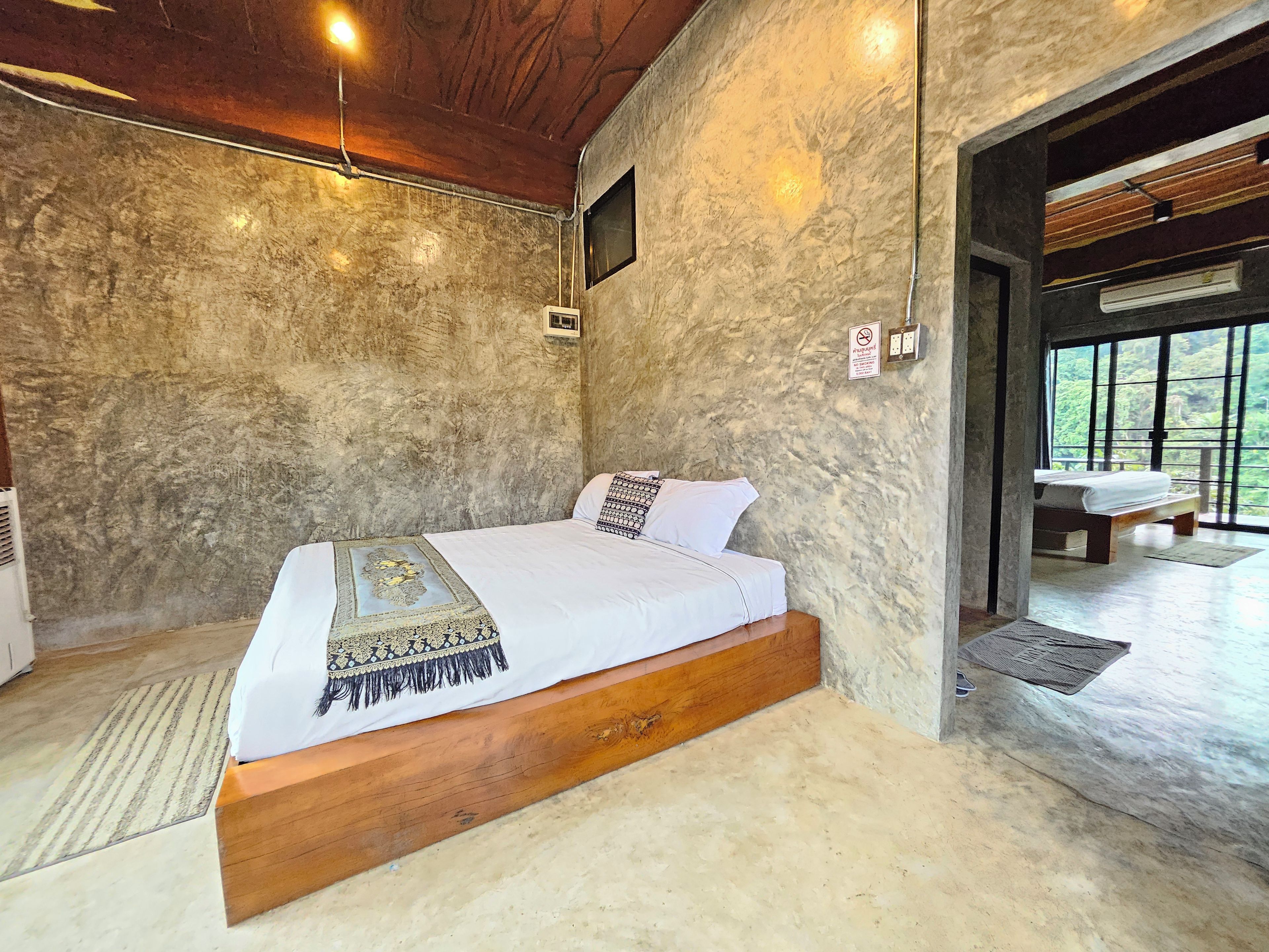 A bedroom with a wooden bed, a stone wall, a small window, and a view of greenery outside.
