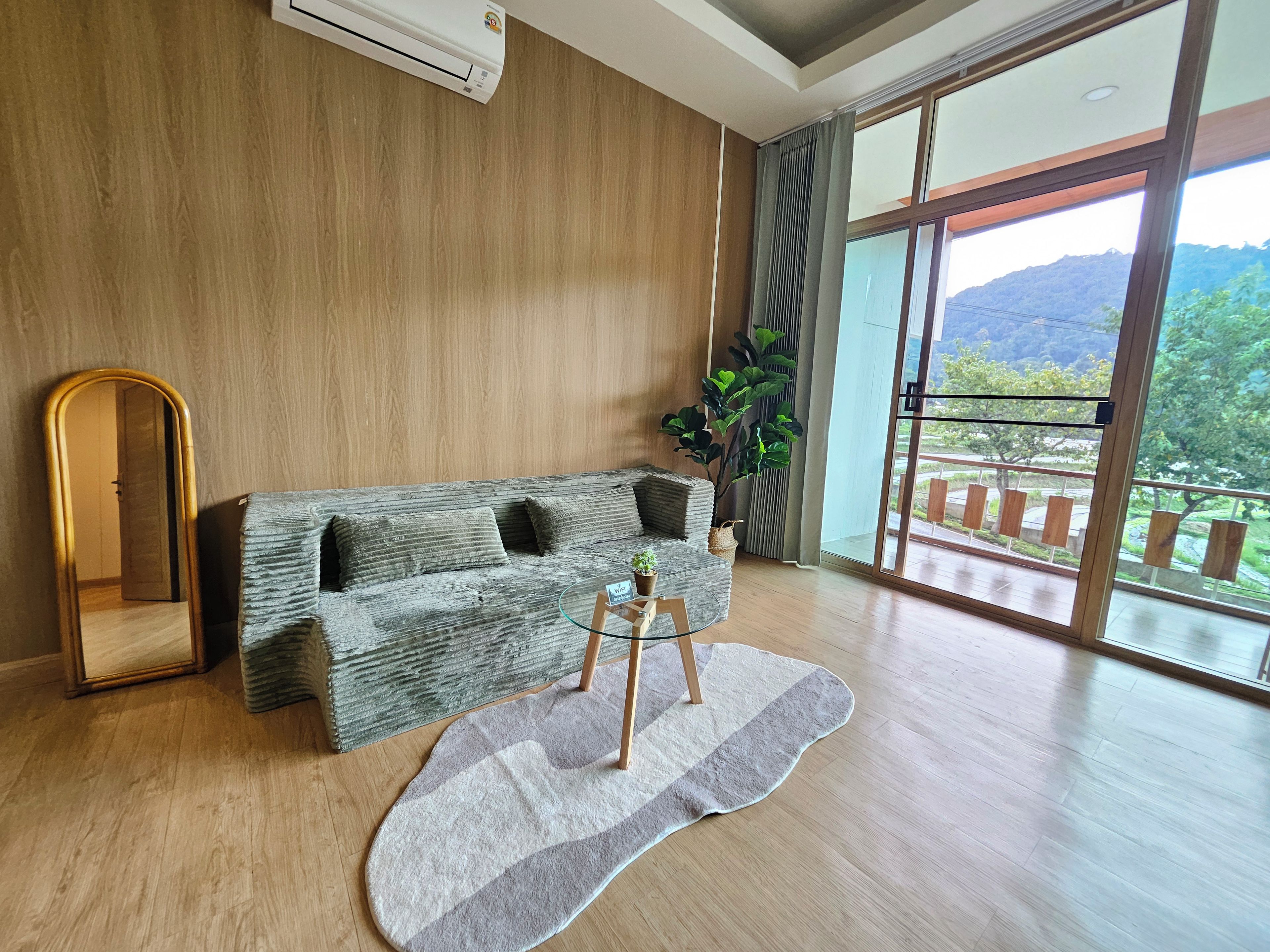 A modern living room with a wooden floor, a grey sofa with cushions, a round coffee table, a mirror, and a large window with a view of greenery and mountains.