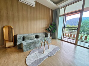 Un salon moderne avec un plancher en bois, un canapé gris agrémenté de coussins, une table basse ronde, un miroir et une grande fenêtre donnant sur un paysage verdoyant et des montagnes.