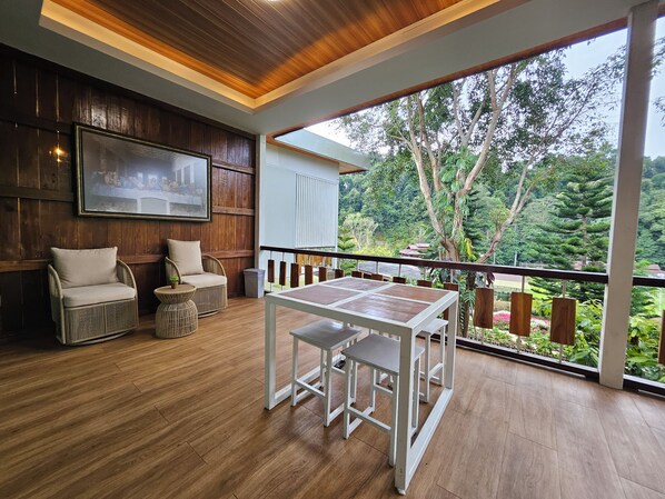 A room with a wooden floor, a dining table with chairs, a sofa, a small round table, and a large window with a view of trees and a house.