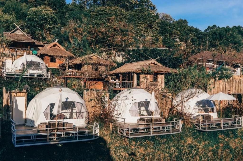 A series of dome-shaped tents with attached wooden decks, surrounded by traditional thatched-roof huts, set against a backdrop of dense forest.