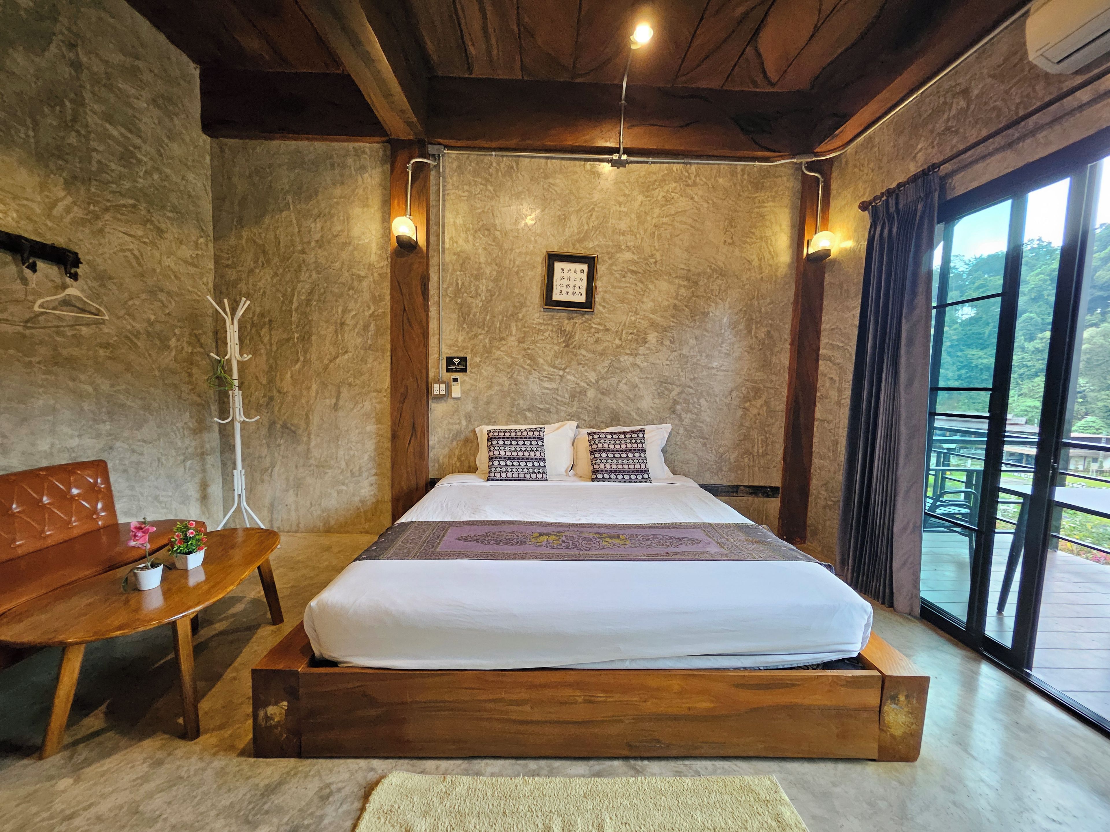 A bedroom with a wooden bed, a bedside table, a coat rack, and a view of the outdoors through a large window.