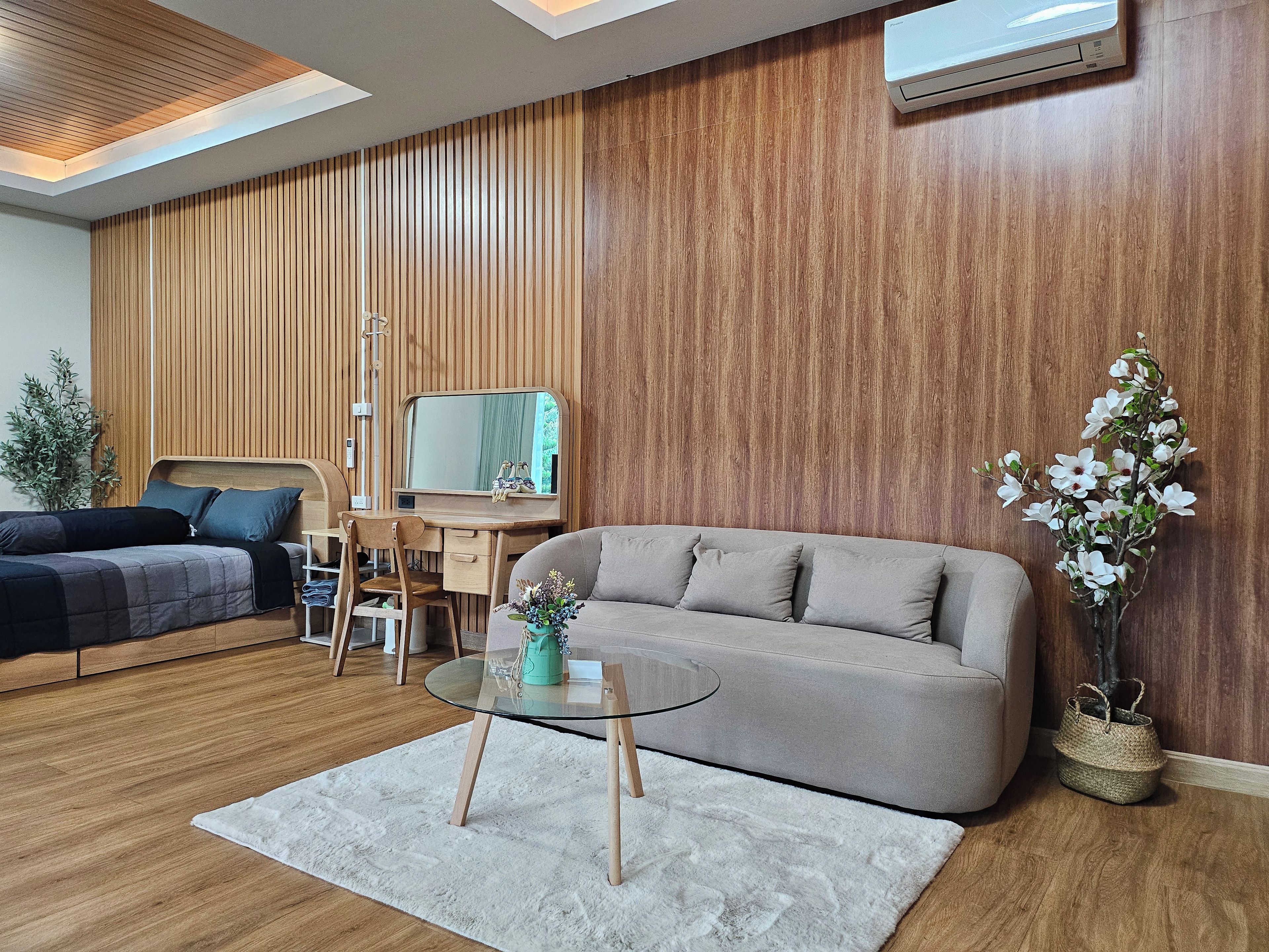 A modern living room with a sofa, a glass coffee table, a desk with a chair, a bed, and a potted plant.