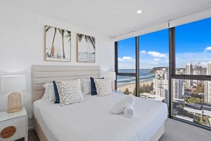 Ein modernes Hotelzimmer mit einem großen Bett, einem Nachttisch, einer Lampe und Blick auf Strand und Stadtlandschaft.