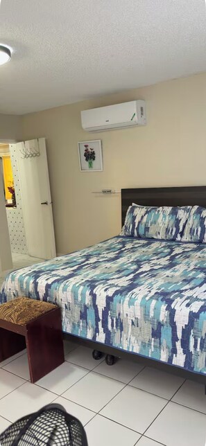 Een slaapkamer met een bed, een klein krukje, een stoel, een airconditioning en een deur.
