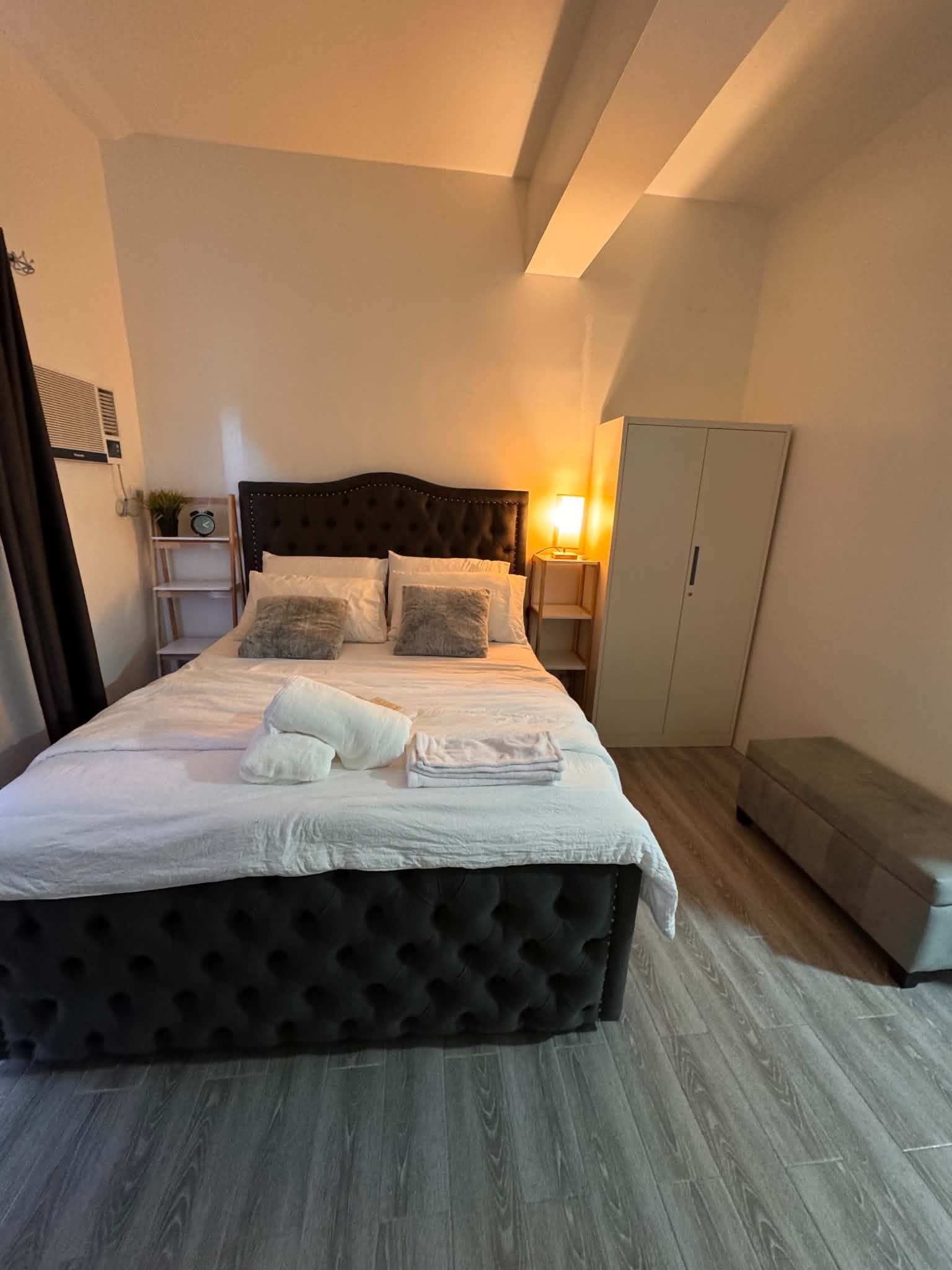 A neatly made bed with a tufted headboard, a bedside table with a lamp, and a wardrobe in a room with wooden flooring.