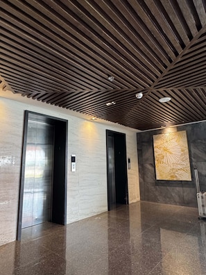 Interior moderno com dois elevadores, piso polido e parede com padrão geométrico.