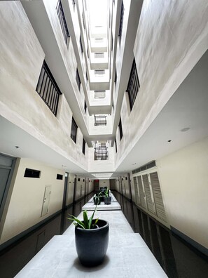 Um corredor com um caminho central, ladeado por paredes com janelas e portas, que leva a um teto alto com claraboia.