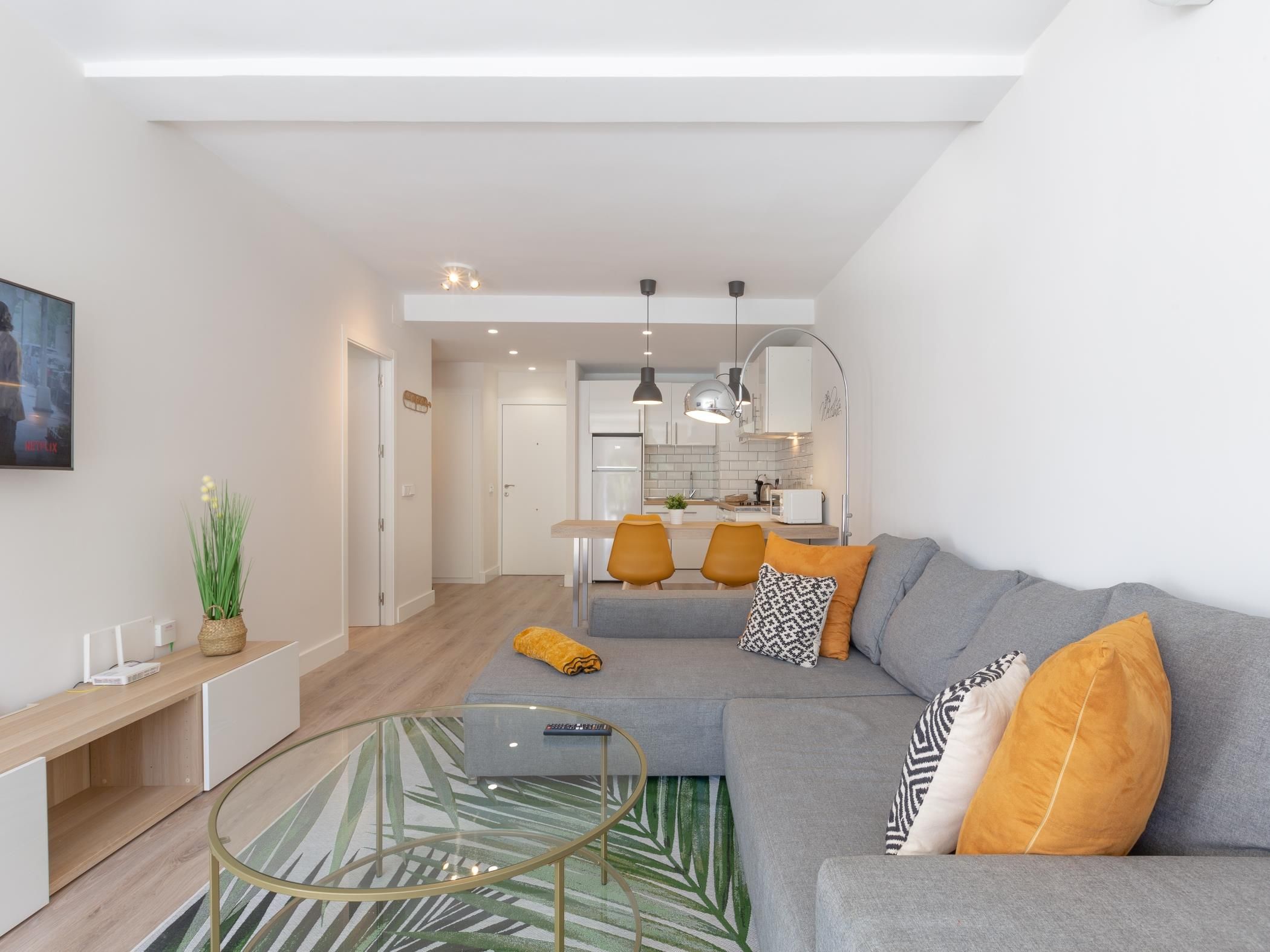 Ein modernes Wohnzimmer mit einem grauen Sofa, einem Glastisch und einer Küche im Hintergrund.