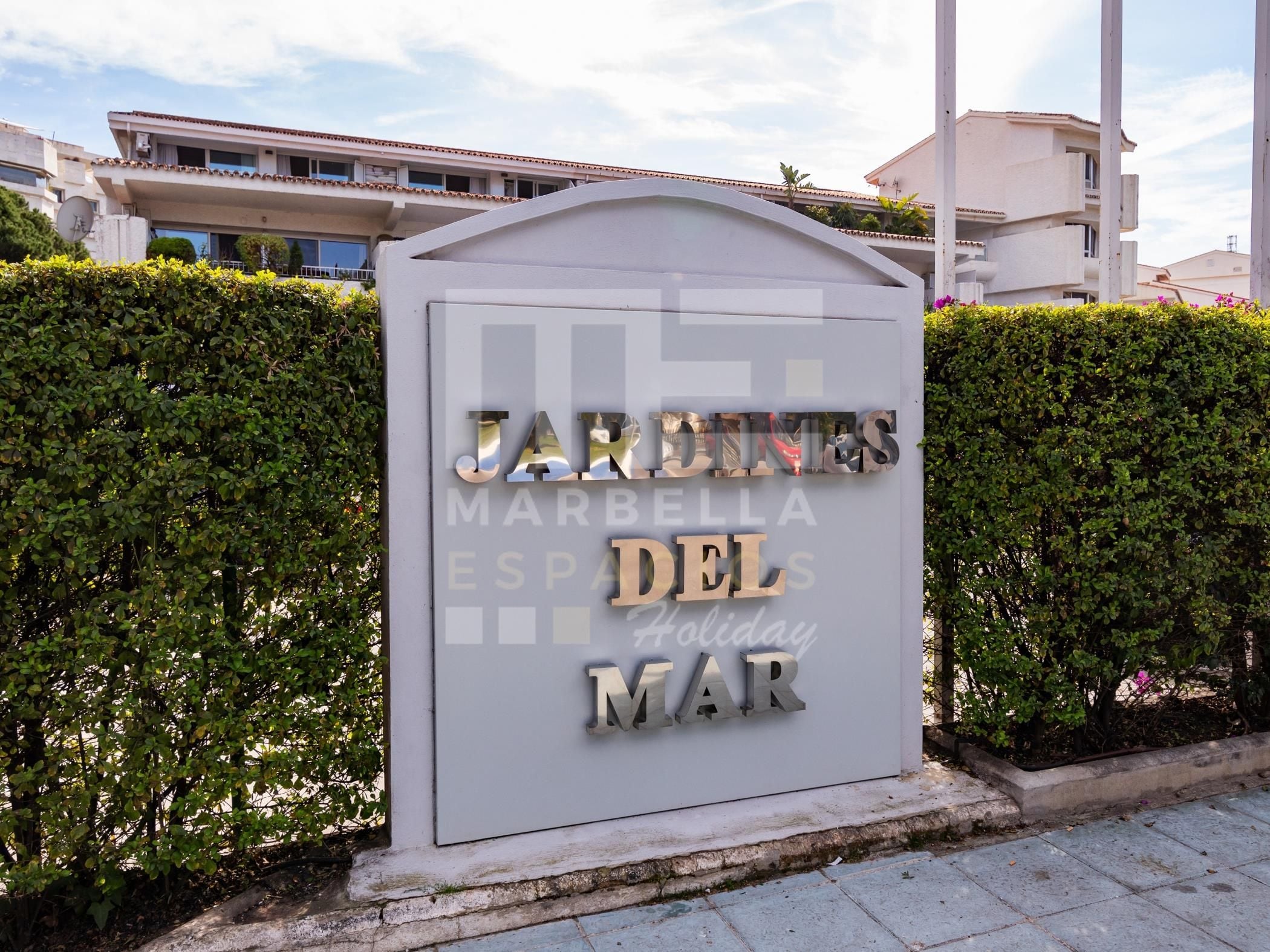 A sign for 'Jardines del Mar' with a modern design, located in front of a building with a tiled roof.