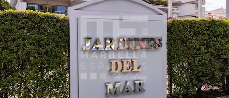 A sign for 'Jardines del Mar' with a modern design, located in front of a building with a tiled roof.