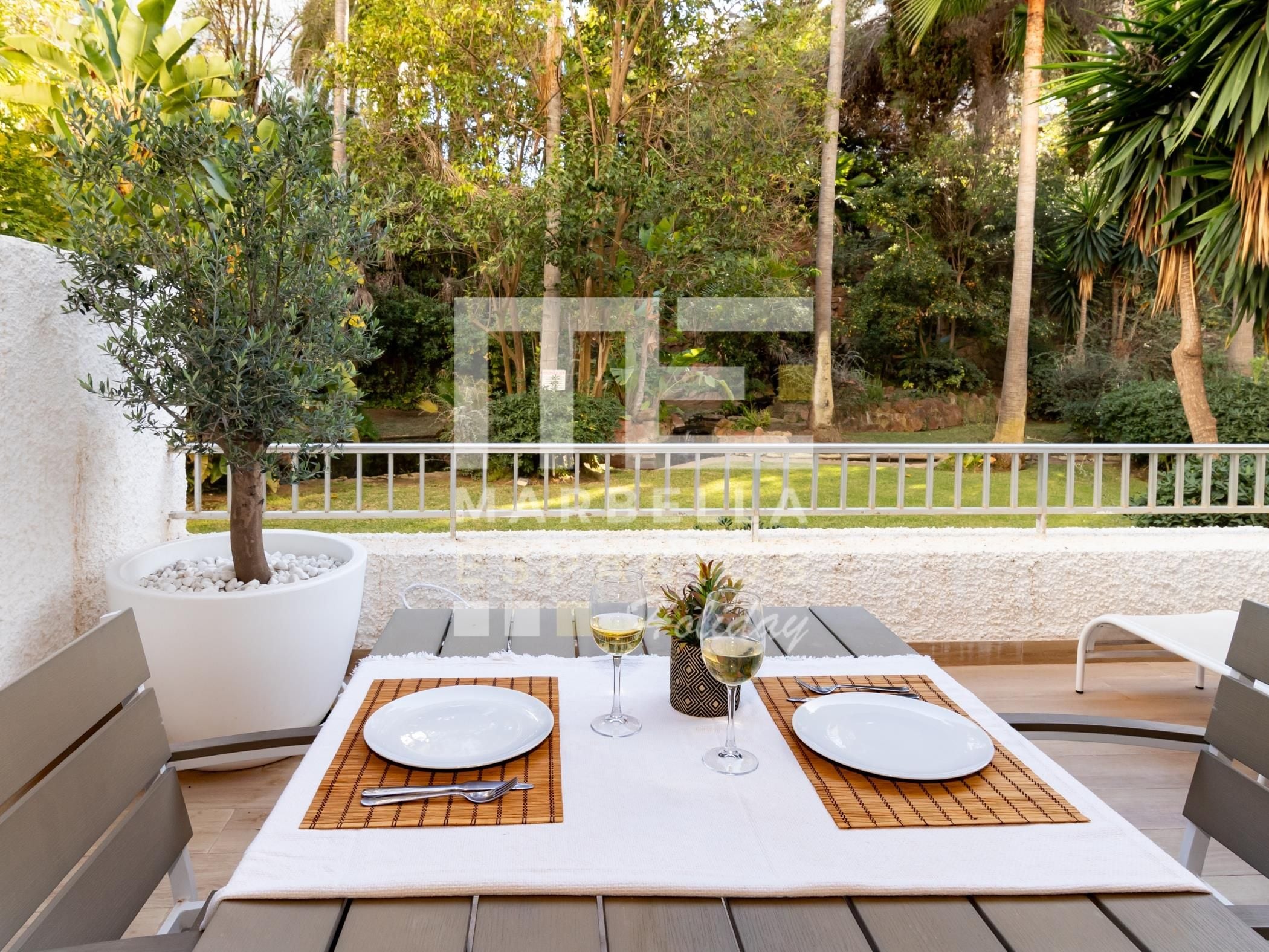 An outdoor dining area with two tables set for two, surrounded by a white railing, with a view of a garden and trees.