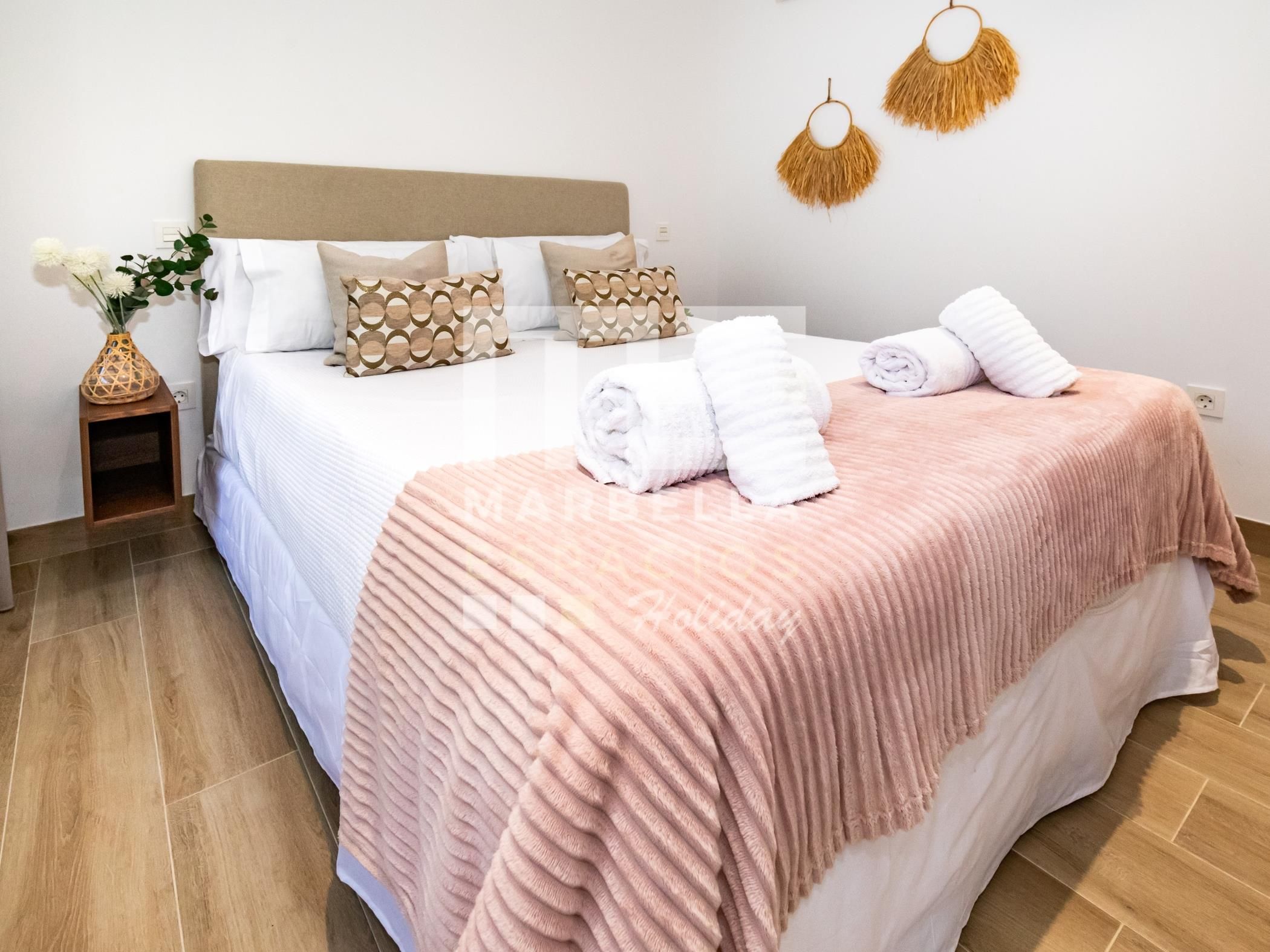 Ein ordentlich bezogenes Bett mit einer rosafarbenen Decke, weißen Kissen und einer Vase mit Blumen auf einem Nachttisch.