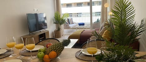 A modern living room with a television, seating area, and a dining table set with fruit and drinks.