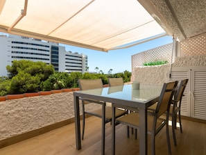 Une terrasse extérieure avec une table et des chaises en verre, un auvent blanc et une vue sur des bâtiments et la verdure.