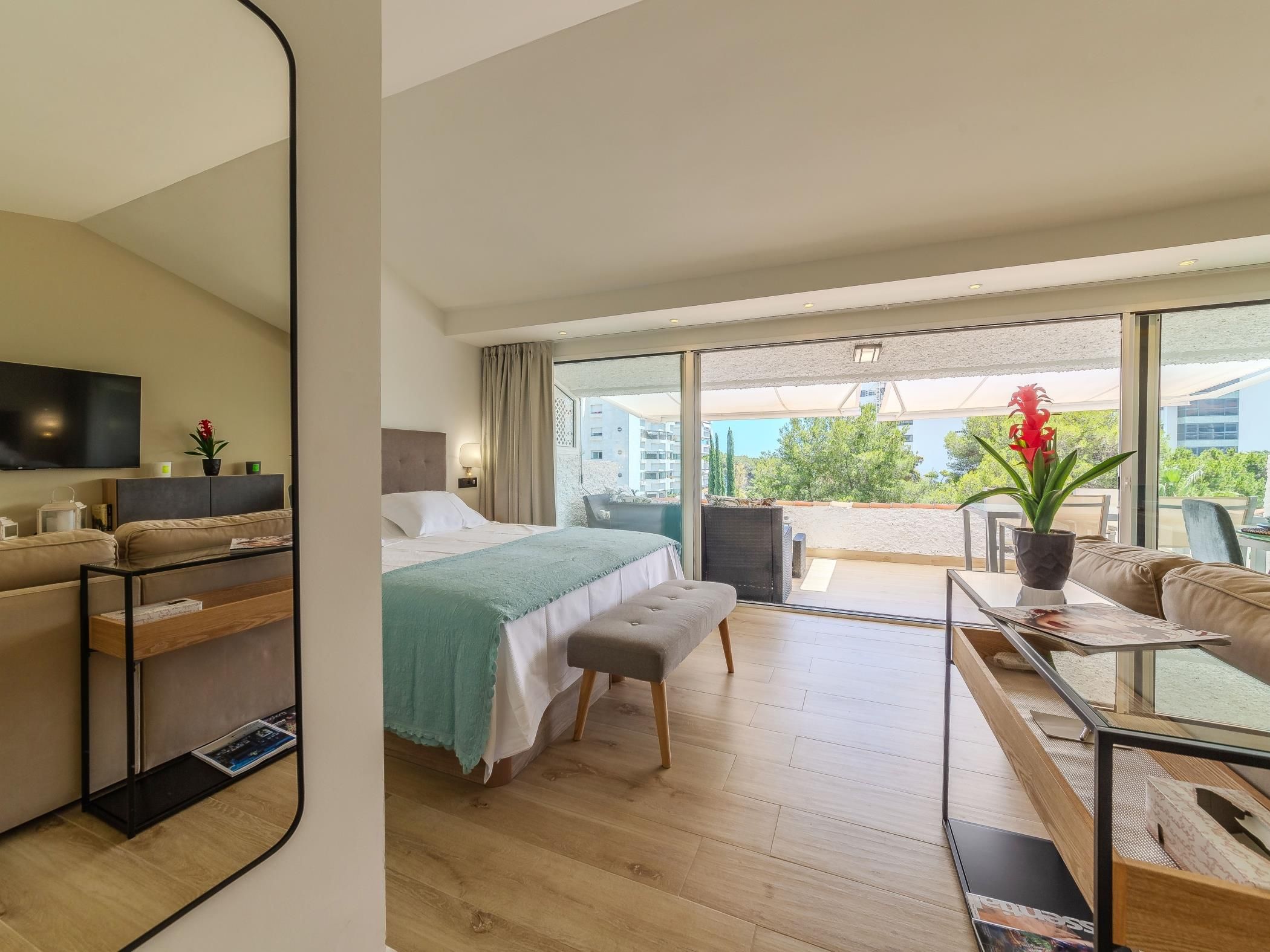Ein modernes Hotelzimmer mit Bett, Sofa, Fernseher und einem Balkon mit Blick auf Gebäude und Grünflächen.