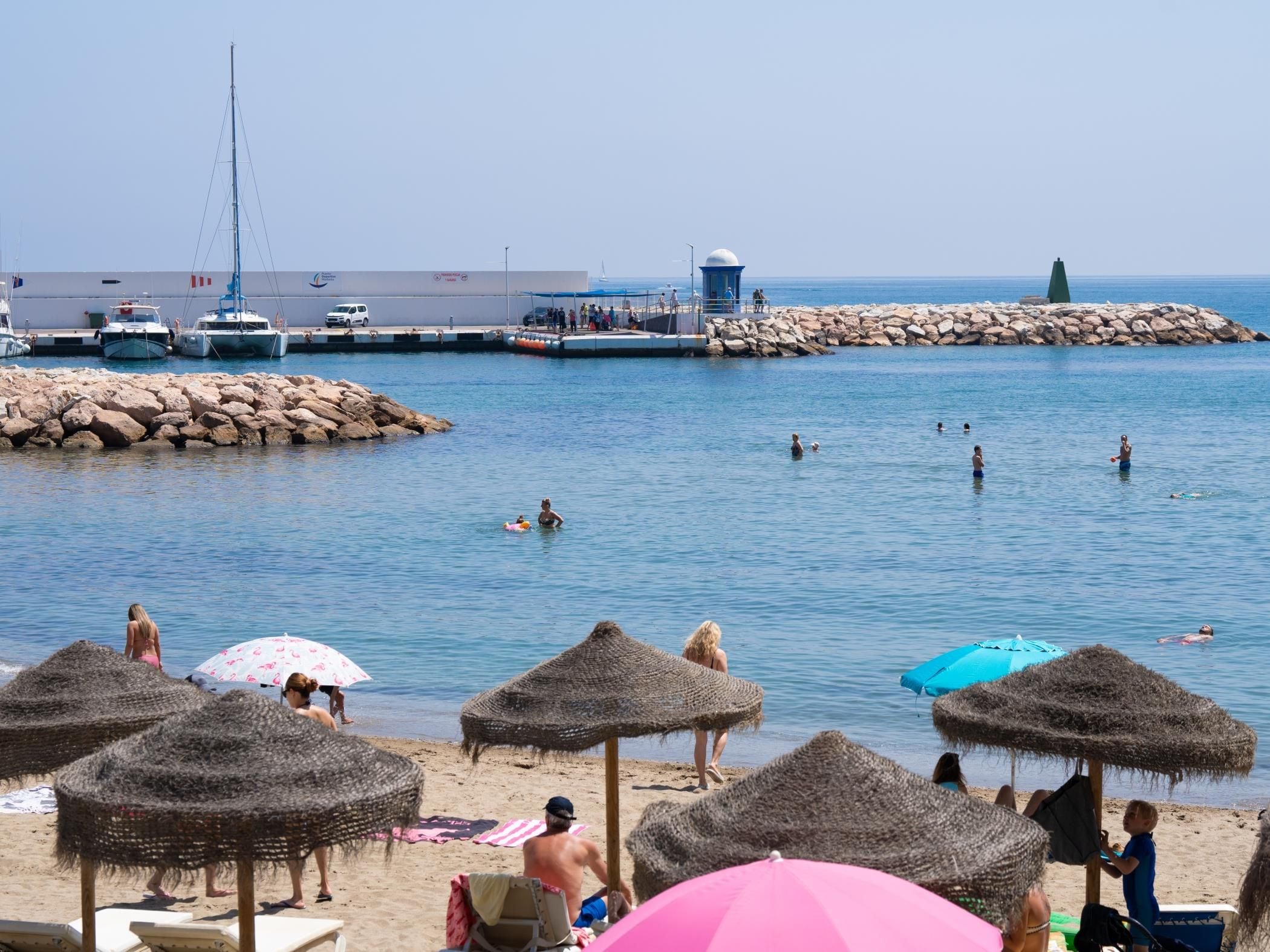 A beach with thatched umbrellas, a rocky breakwater, and boats in the background.