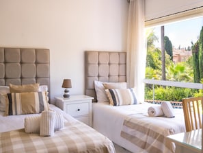 A hotel room with two beds, a nightstand, a lamp, and a view of greenery outside.
