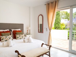 A bedroom with a large bed, a bedside table with a lamp, a mirror, and a view of a garden through a sliding glass door.