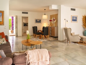 A modern living room with a sofa, coffee table, and chairs. There is a dining area with a table and chairs. The room has a kitchen in the background.