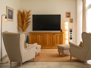 A living room with a large flat-screen TV, wooden cabinet, two armchairs, a floor lamp, and a large plant.