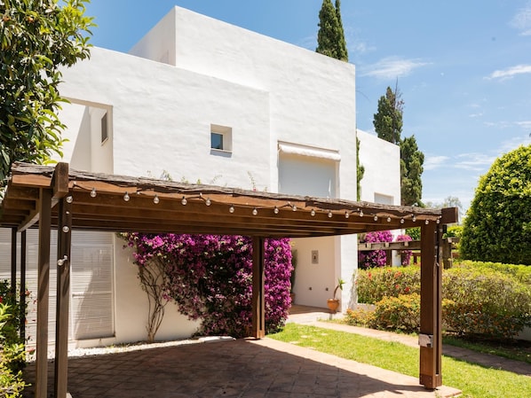A white building with a covered patio area, a pergola, and flowering plants.