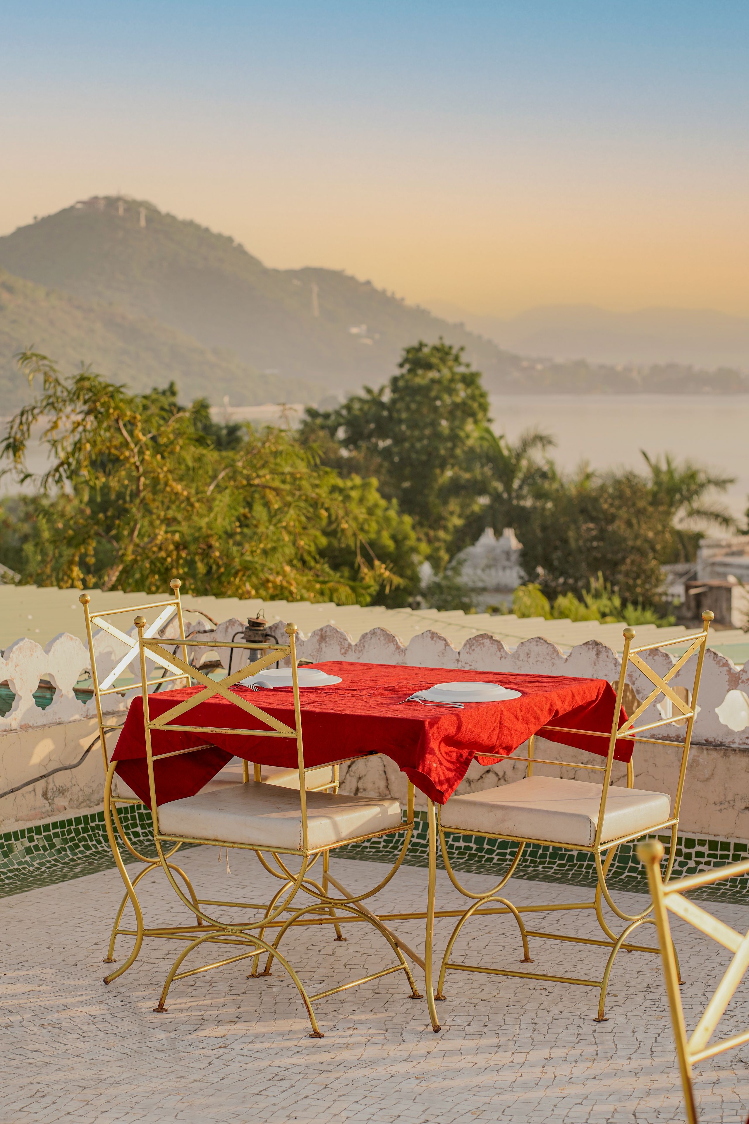 A dining table with a red tablecloth and gold chairs, set on a tiled patio with a view of a distant city and hills.