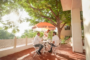 Un grupo de personas sentadas en una mesa bajo una sombrilla, con un árbol y un edificio al fondo.