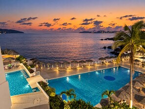 A swimming pool with lounge chairs and umbrellas, overlooking a body of water at sunset. - Beautiful Oceanfront Suite in the Heart of Acapulco (Acapulco de Juárez)