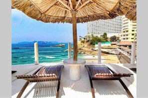 A beachside lounge with a thatched umbrella, lounge chairs, and a view of the ocean and buildings in the background. - Beautiful Oceanfront Suite in the Heart of Acapulco (Acapulco de Juárez)