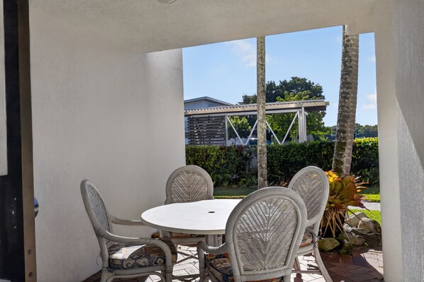 Patio esterno con tavolo rotondo e sedie in vimini, affacciato su un giardino con una pergola e una casa sullo sfondo.