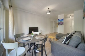 A modern living room with a grey sofa, a dining table set with plates, and a television in the background. - 14522433 FRGK Stanislas Apartment Terrace, Central (Cannes)