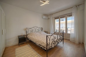A bedroom with a metal bed frame, a bedside table, a nightstand, a window with curtains, and a view of buildings outside. - 14522433 FRGK Stanislas Apartment Terrace, Central (Cannes)