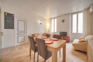 Uma sala de jantar com mesa posta para quatro pessoas, um sofá, uma TV e uma janela com cortinas.
