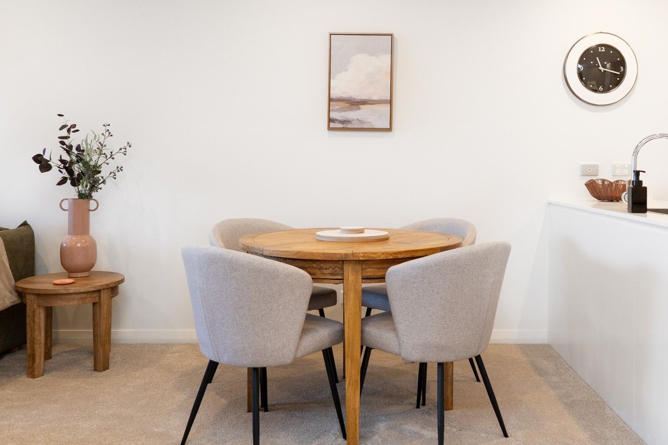 A dining area with a round wooden table, two grey chairs, a vase with plants, a painting on the wall, and a clock.