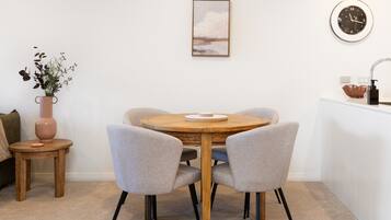 A dining area with a round wooden table, two grey chairs, a vase with plants, a painting on the wall, and a clock.